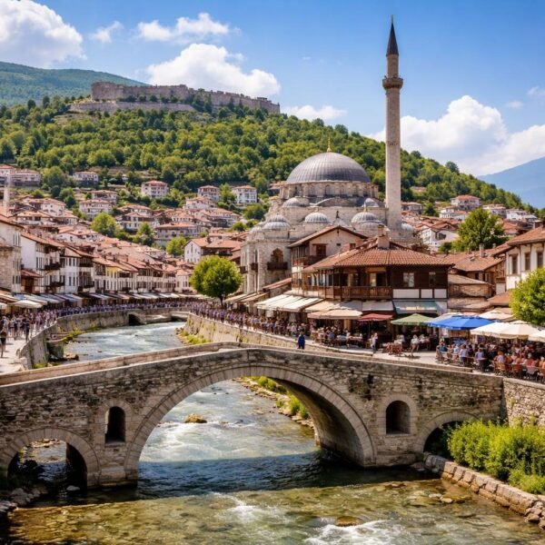 Vista da cidade de Prizren com pontes de pedra, arquitetura tradicional, pessoas caminhando e a fortaleza ao fundo sob céu azul.